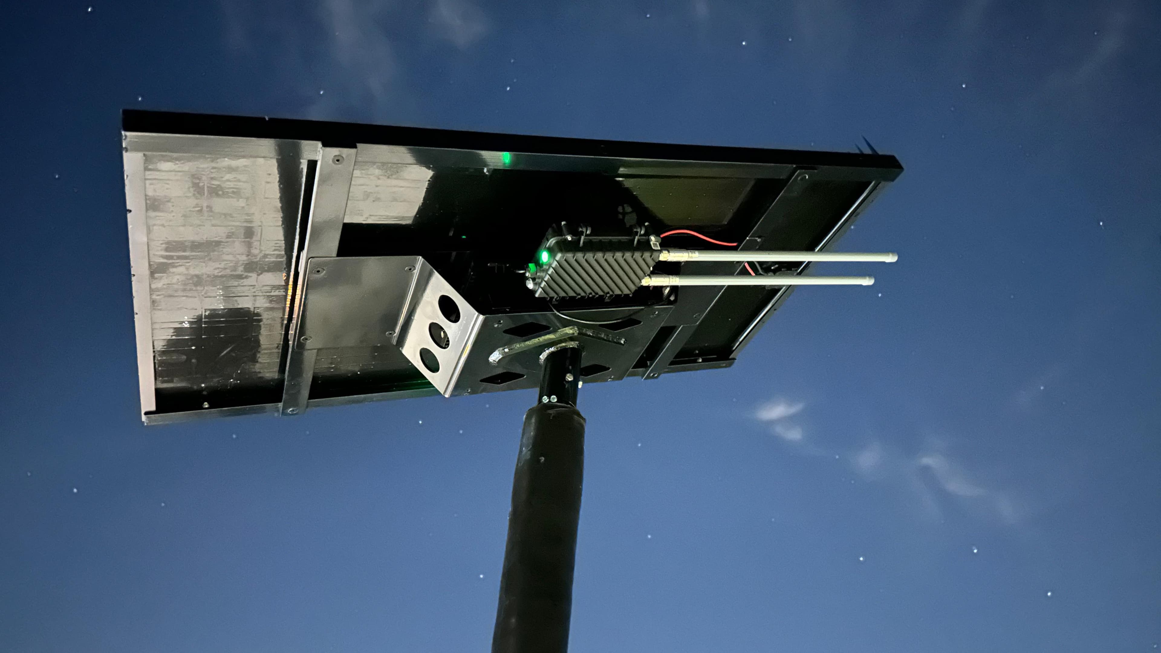 Underside detail of the All Seasons pole top at night, showing the panel, light head, cellular and Wi-Fi antennas, and a green status LED